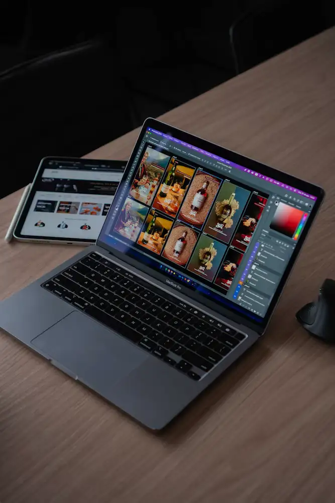 Notebook aberto em um programa de edição de imagens, com um tablet mostrando uma loja online ao lado, sobre uma mesa de madeira