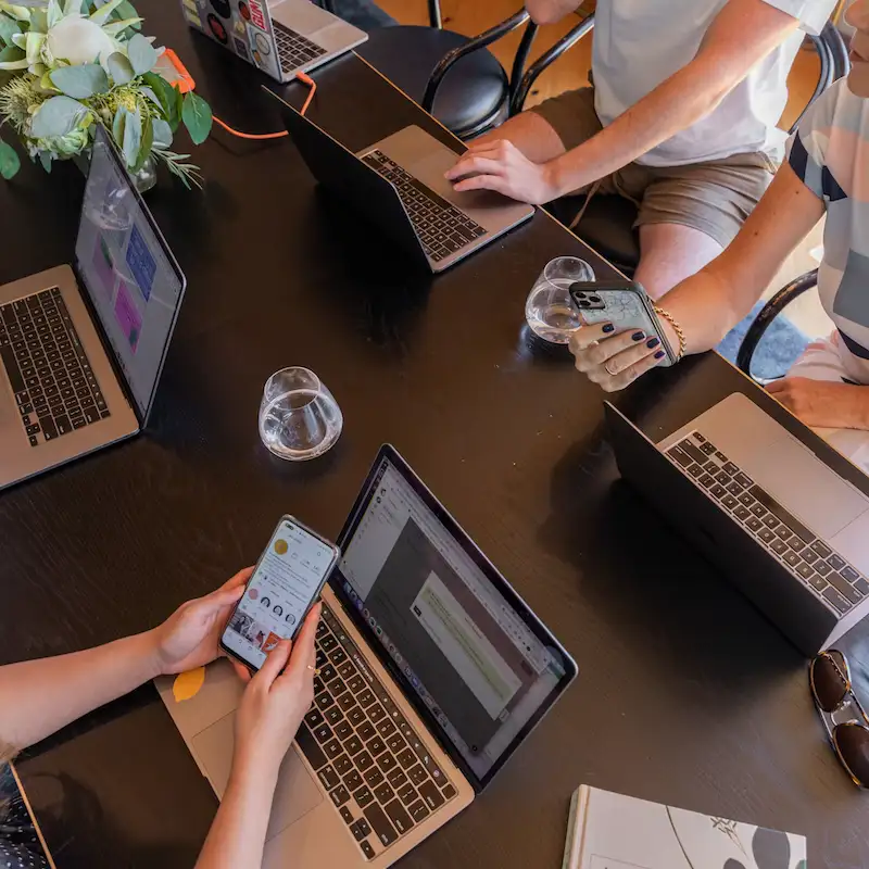 Vista de cima de pessoas trabalhando em notebooks ao redor de uma mesa escura, uma delas mexendo no celular