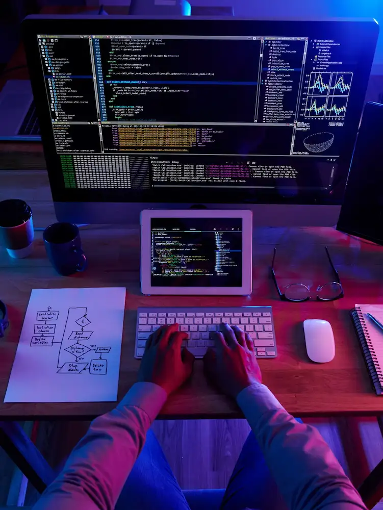 Pessoa digitando diante de um monitor com código, com um tablet e um fluxograma desenhado em papel sobre a mesa