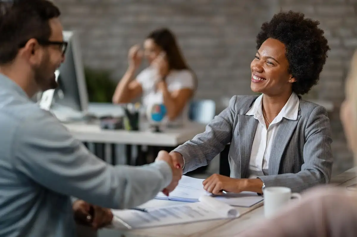 Mulher de blazer cinza sorrindo e apertando a mão de um cliente sobre uma mesa com papéis, em um escritório