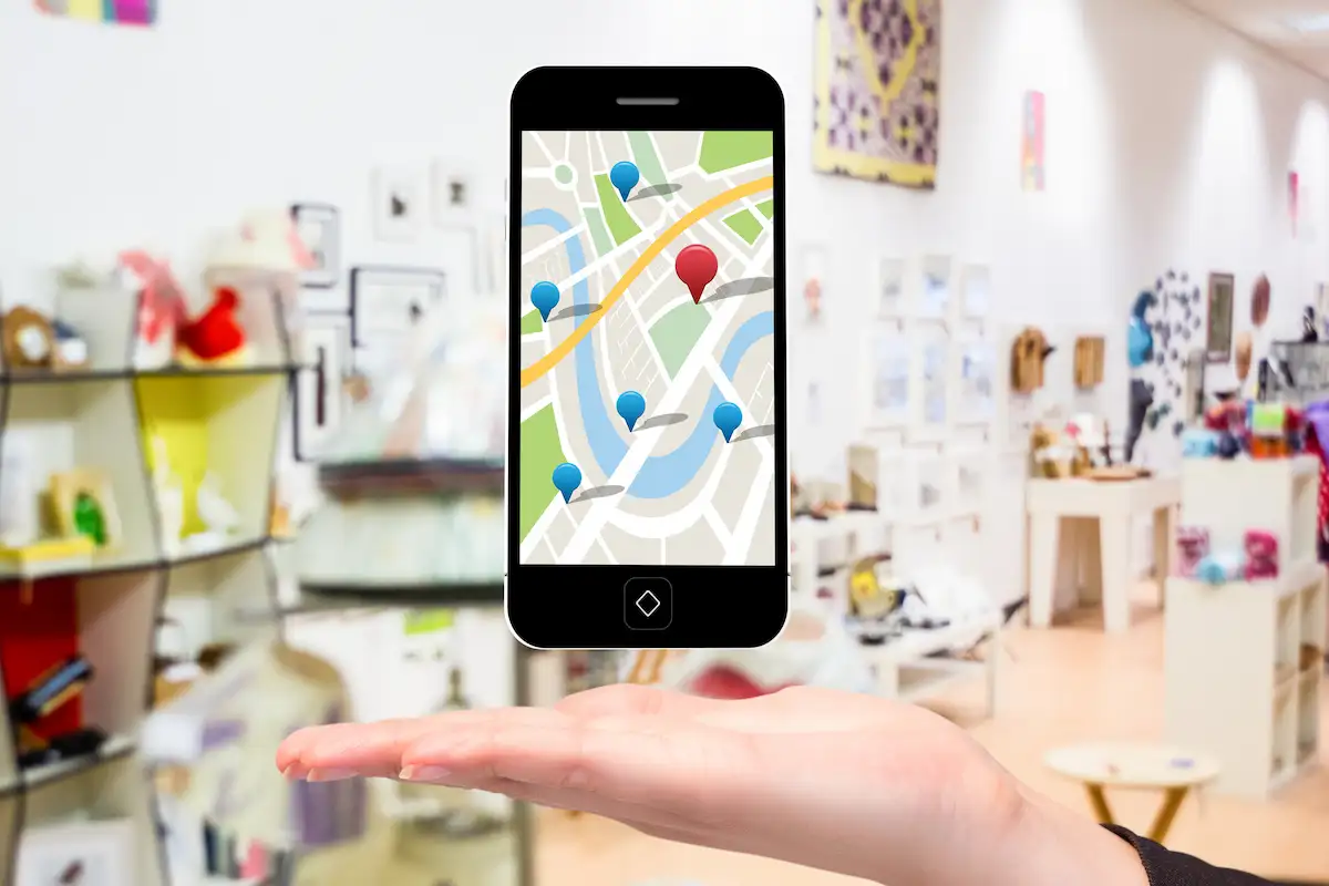 Celular com um mapa e marcadores de localização flutuando sobre a palma de uma mão, dentro de uma loja com prateleiras ao fundo