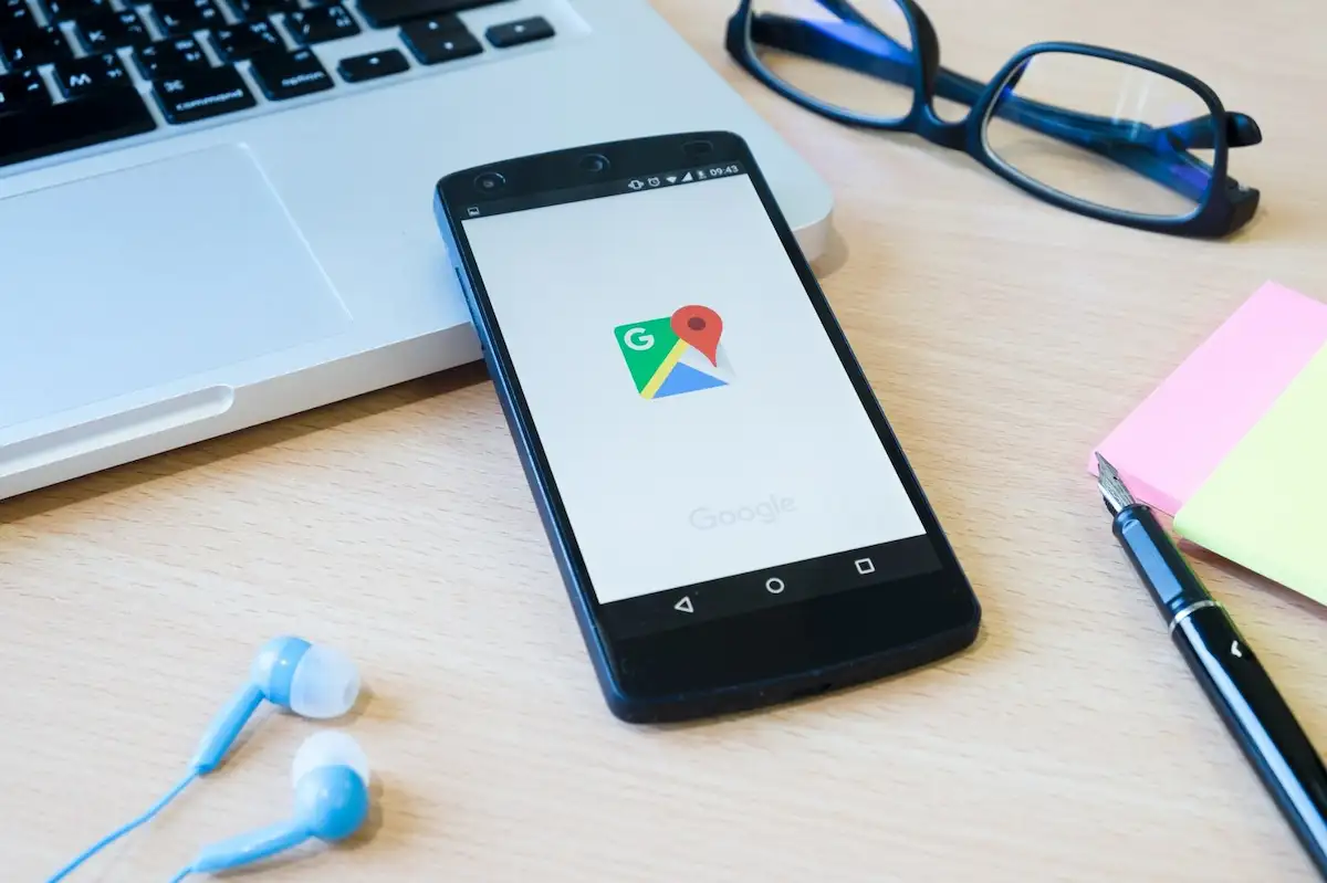 Celular com o ícone do Google Maps na tela, sobre uma mesa ao lado de um notebook, óculos, fones de ouvido, caneta e blocos de anotação