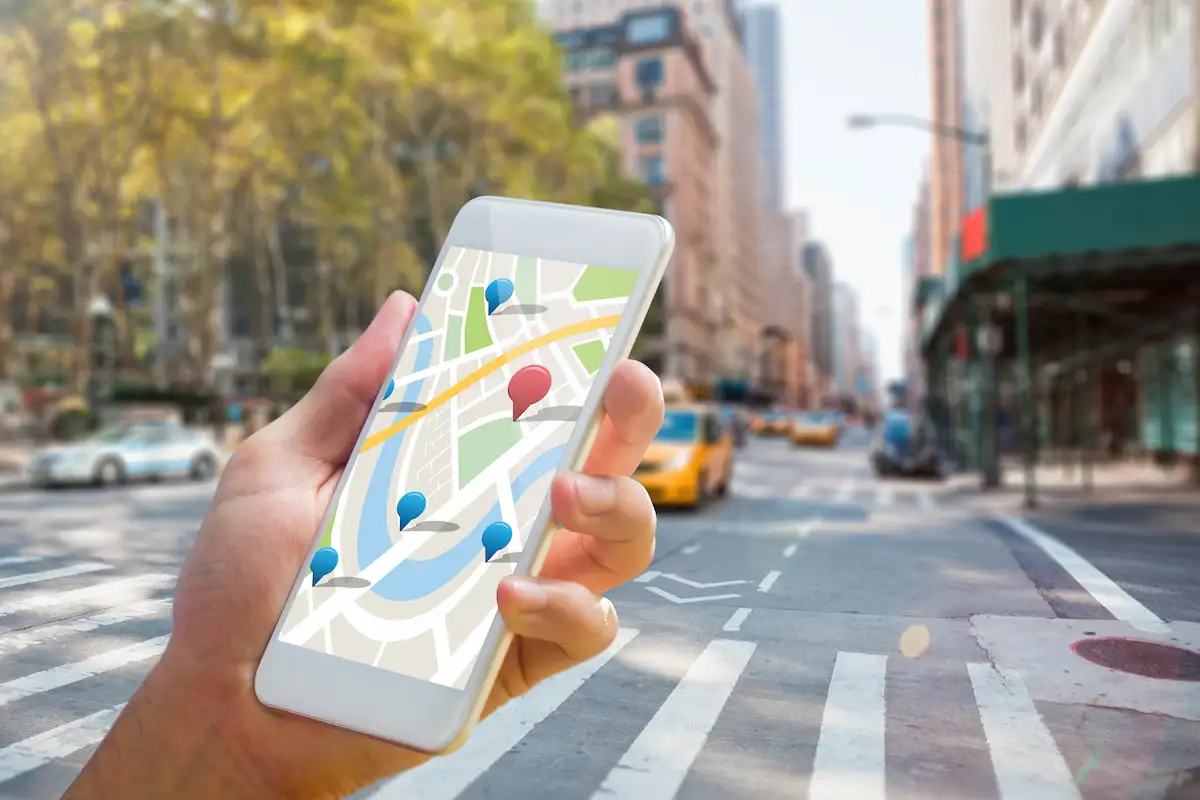 Mão segurando um celular com um mapa e marcadores de localização na tela, numa rua de cidade com táxis amarelos e prédios ao fundo