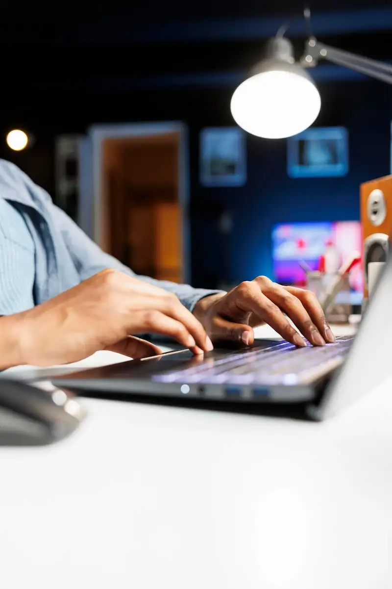 Mãos digitando em um notebook sobre uma mesa branca, em um escritório com luz baixa