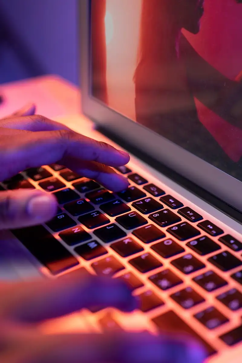 Mãos digitando em um teclado de notebook iluminado em tons de rosa e azul