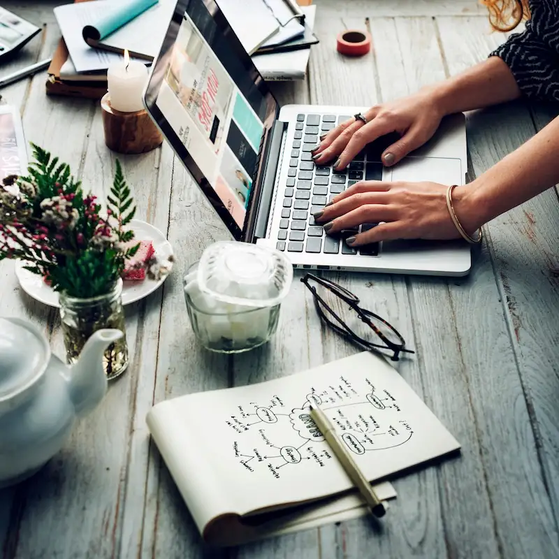 Mãos digitando em um notebook sobre uma mesa de madeira, ao lado de um caderno com esboços de fluxo