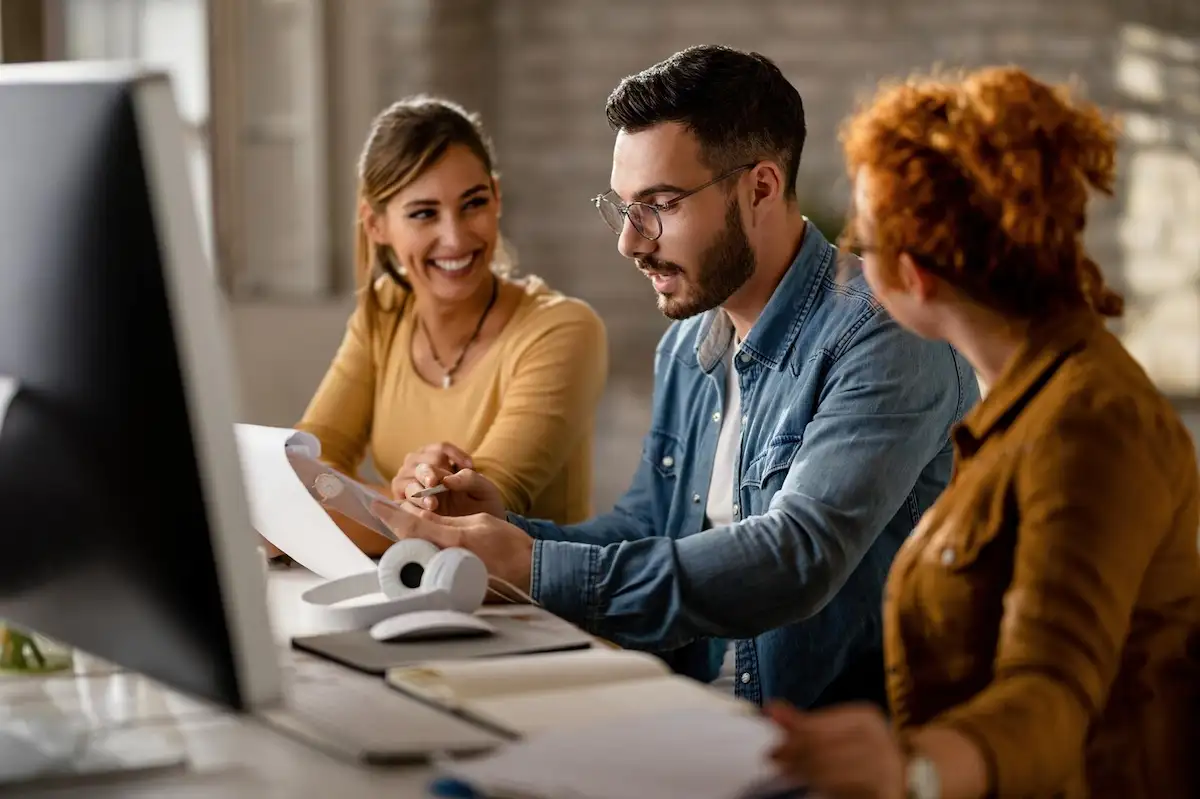 Três pessoas conversando sorridentes diante de um computador, uma delas segurando papéis