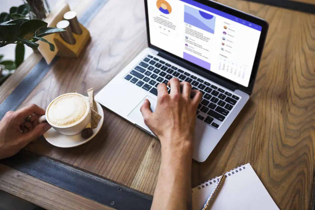 Mão digitando em um notebook com um painel de gráficos na tela, ao lado de uma xícara de café