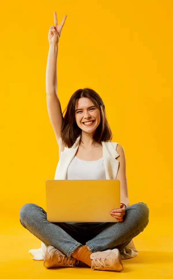 Mulher sorridente sentada no chão com um notebook no colo, fazendo sinal de vitória sobre fundo amarelo