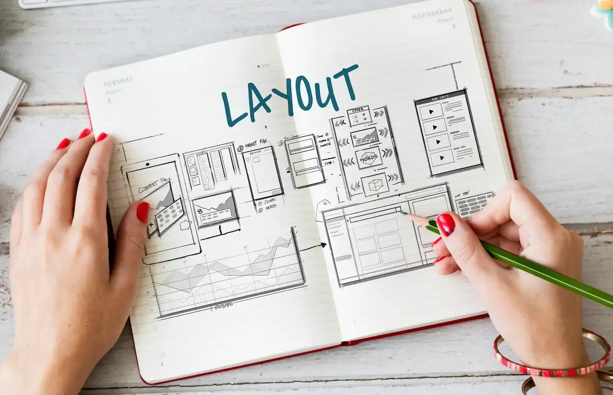 Caderno aberto com esboços de páginas de site desenhados a lápis e a palavra LAYOUT no alto, com uma mão segurando o lápis