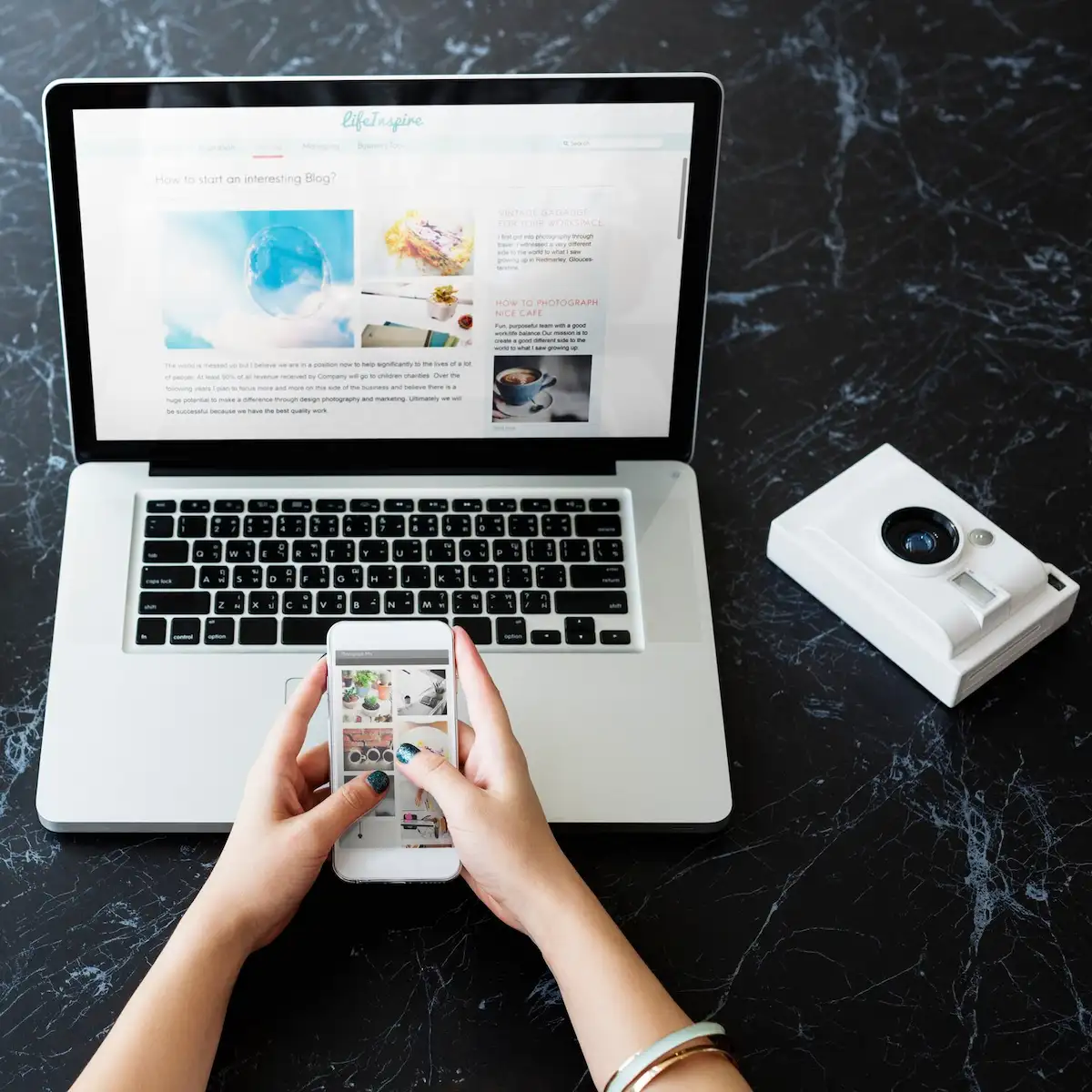 Mãos segurando um celular diante de um notebook com um blog aberto na tela, ao lado de uma câmera instantânea branca, sobre uma mesa de mármore preto