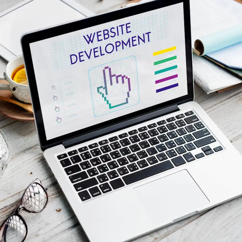Notebook com a frase Website Development e o desenho de uma mão clicando na tela, ao lado de óculos e de uma xícara de chá