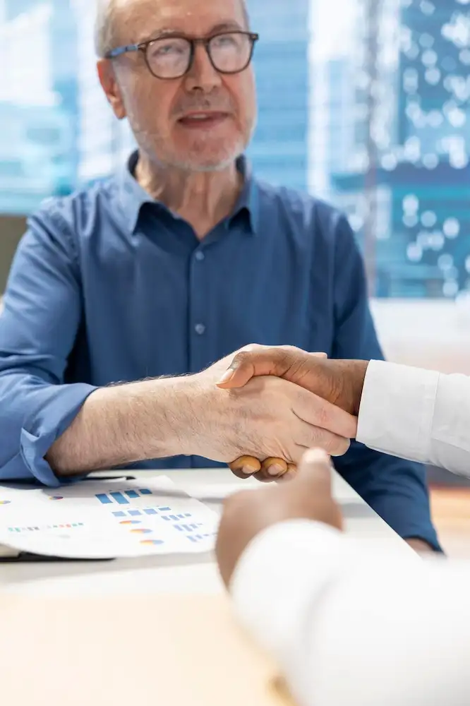 Homem de óculos e camisa azul apertando a mão de outra pessoa sobre uma mesa com gráficos impressos